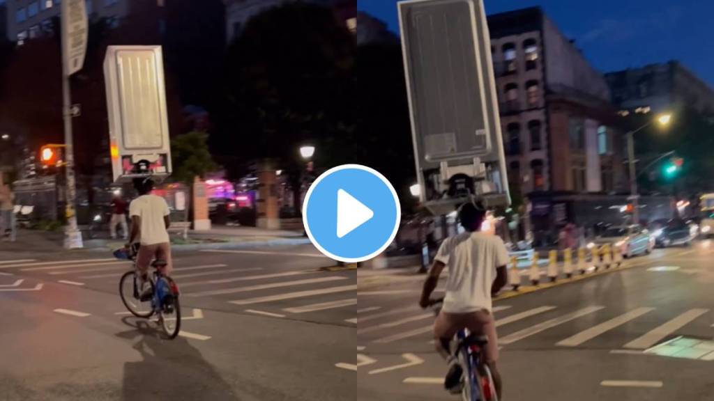 A person is seen riding a bicycle on the road with a fridge on his head