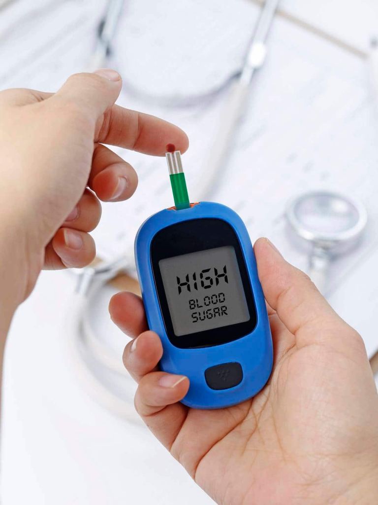 Hand holding a blood glucose meter measuring blood sugar, the background is a stethoscope and chart file